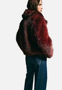 Czerwona sztuczna futrzana kurtka z wysokim kołnierzem, luźnym krojem i teksturowanym wykończeniem. Połączona z ciemnoniebieskimi dżinsami, zaprezentowana w widoku trzech czwartych.