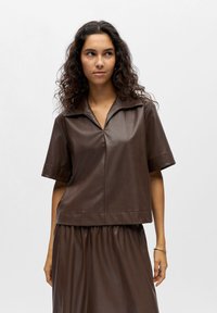 Femme aux longs cheveux bouclés portant un haut en cuir marron foncé à manches courtes et une jupe assortie, debout devant un fond clair uni.