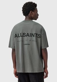 Camiseta de gran tamaño y manga corta en algodón verde apagado, con texto negro "ALLSAINTS" y "LONDRES" en la parte posterior. Textura suave.