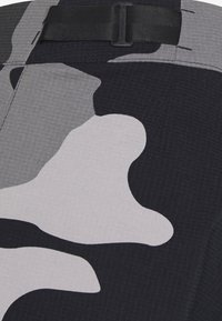 Kamouflage-tyg med abstrakta former i grått, vitt och svart. Har en texturerad yta och en justerbar svart remdetalj.