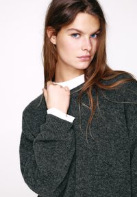 Nadrozmerný sveter tmavosivej farby s textúrovaným pletením a širokými manžetami, vrstvený cez bielu košeľu s golierom, zachytávajúci jeho pohodlný strih.