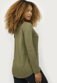 Frau mit lockigem Haar, die über die Schulter schaut, trägt ein olivgrünes Langarmshirt und schwarze Hosen vor einem weißen Hintergrund.