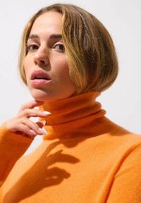 Nahaufnahme einer Person, die einen gerippten orangefarbenen Rollkragenpullover trägt. Der Hintergrund ist weiß, was die leuchtende Farbe und Textur hervorhebt.