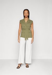 Blazer senza maniche verde oliva con design doppiopetto, vita cintata e due tasche frontali; abbinato a pantaloni bianchi a gamba larga e tacchi neri.