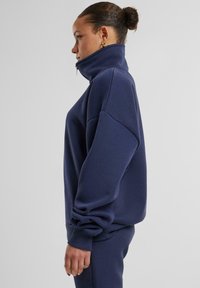 Felpa blu navy con collo alto e dettaglio con zip, spalle scese e polsini a coste. Realizzata in tessuto morbido e elasticizzato.