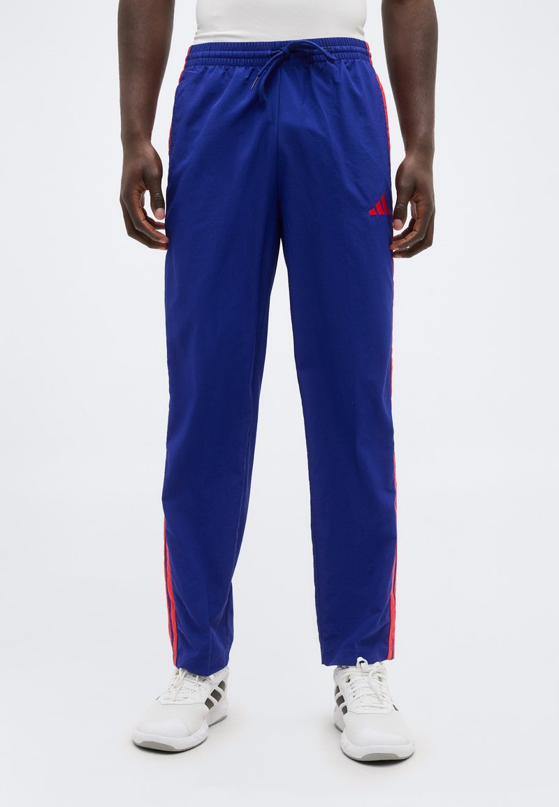 Mężczyzna ubrany w niebieskie spodnie treningowe Adidas z czerwonymi paskami i białe trampki, stojący na tle białego tła.