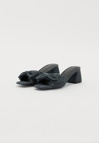 Mule in denim scuro con tacco alto, punta aperta e un grande fiocco davanti. Tacco a blocco arrotondato e texture morbida.