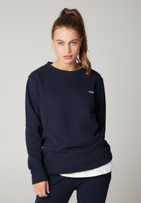 Felpa blu navy in misto cotone, con maniche lunghe, scollo tondo e un piccolo emblema sul petto. Texture morbida.
