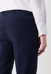 Mørkeblå chinos laget av en bomullsblanding med sydde sidelommer og baklommer. Har slim fit og glatt tekstur, kombinert med en hvit skjorte.