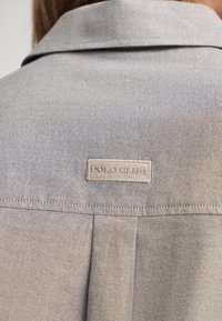 Ljust grå denimskjorta med krage och en signaturtagg "POLO CLUB" på baksidan, med synliga sömnadsdetaljer.