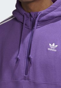 Lila hoodie med dragkedja framtill, snören och vitt Adidas-logotyp. Har texturerad tyg och kontrasterande ränder på ärmarna.