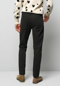 Dunkle, schmal geschnittene Hose mit Gesäßtaschen, aus einem glatten Stoff. Dazu wird ein gemusterter Pullover und braune Stiefel getragen.