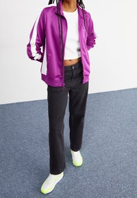 Lila zip-up-jacka med vita ränder, kombinerad med en vit croppad topp, svarta jeans och sneakers med gröna detaljer på en texturerad blå yta.