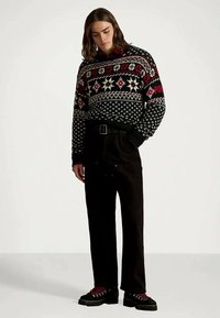 Młoda osoba z włosami do ramion nosi sweter w czarno-czerwono-białe wzory, czarne spodnie i czarne buty z czerwonymi sznurowadłami, stoi.