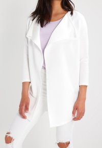 Bílý cardigan s jemnou texturou, otevřený vpředu a s volným límcem, spárovaný s levandulovým topem a roztrhanými bílými džíny.