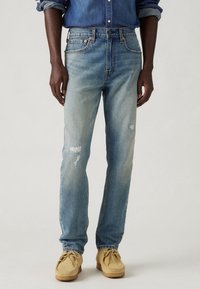 Světle modré džíny z denimu s vybledlými oblastmi a mírnými oděry. Mají klasický styl s pěti kapsami a rovný střih nohavic. K džínsům jsou nošeny béžové mokasíny.