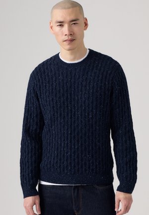Homme portant un pull en maille torsadée bleu marine par-dessus une chemise blanche et un jean foncé sur un fond uni gris clair.