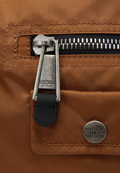 Brown nylon bag detail featuring a silver zipper, black leather zipper pull, and a metallic Moschino logo button. Stitched edges are visible.