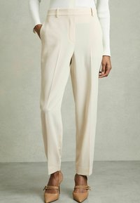 Pantaloni beige su misura con tasche laterali, frontale plissettato e gamba affusolata, abbinati a décolleté nude con punta su uno sfondo neutro.