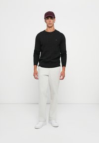 Άνδρας που στέκεται φορώντας ένα μαύρο πλεκτό πουλόβερ, ανοιχτό γκρι παντελόνι, λευκά αθλητικά παπούτσια και σκούρο κόκκινο καπέλο μπέιζμπολ, με φόντο λευκό.