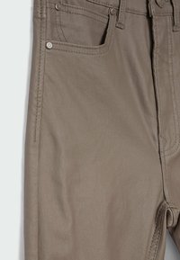 Gros plan sur un pantalon taupe montrant la poche avant, les rivets en métal, les coutures et les passants de ceinture.