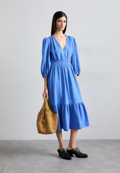 Blauwe, knielange jurk met opgeblazen mouwen, textuurstof en gerimpelde taille. Bruine suède tas en zwarte hakken met decoratieve accenten.