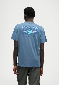 T-shirt de algodão azul com um grande logótipo refletor "Rip Curl" e um gráfico do oceano nas costas; mangas curtas e um corte descontraído.