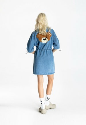 Mujer con cabello rubio que lleva un vestido vaquero azul con un diseño de oso de peluche en la espalda, calcetines blancos con diseño negro y zapatos beige.