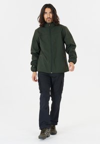 Veste softshell vert foncé avec fermeture éclair à l'avant, poches latérales et poignets élastiques, associée à un pantalon cargo noir et des bottes de randonnée marron.