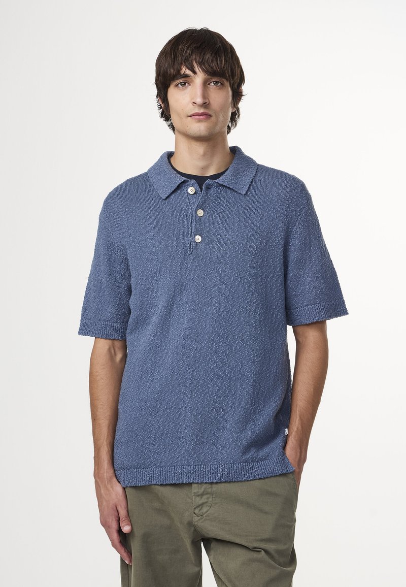 Polo a maniche corte in maglia azzurra chiara con texture, dotato di colletto, patta con tre bottoni e orlo dritto. Abbinato a pantaloni verdi.