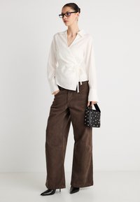 Λευκό πουκάμισο τύπου wrap με μακριά μανίκια και ζωνάκι στη μέση, σε συνδυασμό με χαλαρά, καφέ παντελόνια με ψηλή μέση. Μαύρη τσάντα με τρουκς και γόβες με μυτερή μύτη.