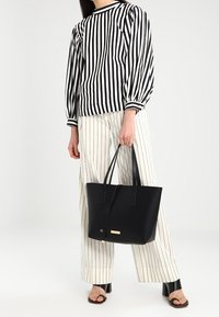 Sac cabas noir à texture lisse, design structuré et deux poignées. Présente un petit accent doré à l'avant. Chemisier rayé et pantalon portés.