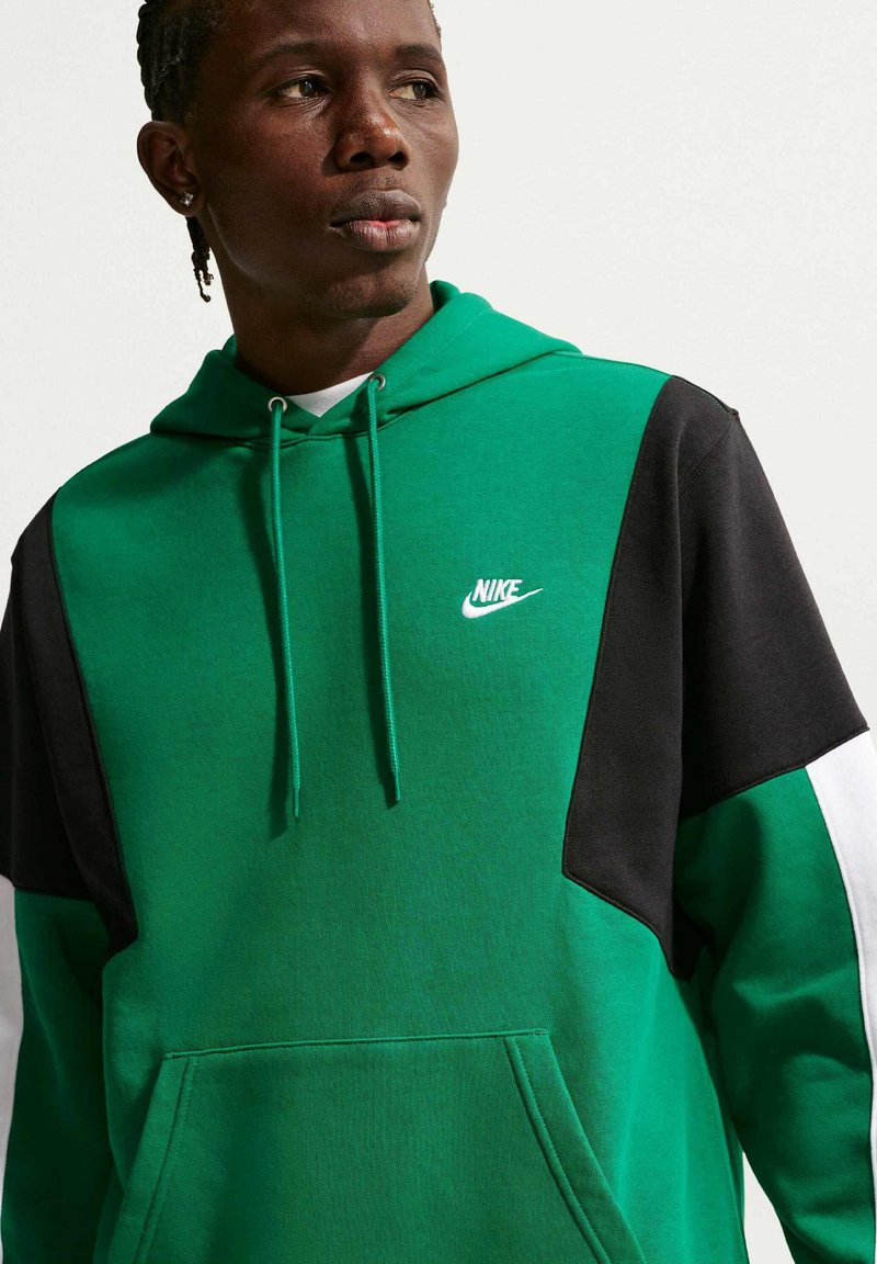 Giovane uomo con trecce corte che indossa una felpa con cappuccio Nike verde, nera e bianca, che guarda di lato su uno sfondo semplice.