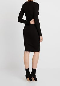 Schwarzes, geripptes Langarmkleid, figurbetont, knielang, mit rundem Ausschnitt. Das Modell trägt schwarze, Absatzstiefel bis zum Knöchel.