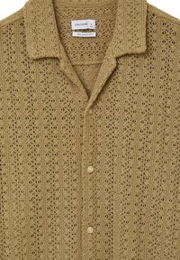 Camicia button-up in maglia all'uncinetto beige con una vestibilità rilassata, motivo a trama aperta, colletto corto e tre bottoni visibili sulla parte anteriore.