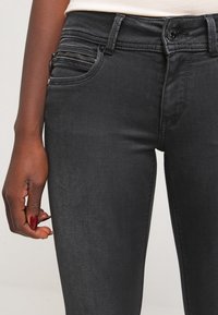Jeans de mezclilla negra con un diseño ajustado, que presentan un detalle de bolsillo con cremallera en el lado izquierdo y una textura suave.