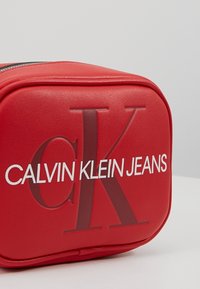 �Červená obdélníková taštička se zipem, se zvýrazněným logem "cK" a bílým textem "CALVIN KLEIN JEANS" na přední straně.