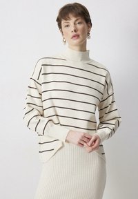 Pruhovaný krémový sveter s hnedými pruhmi, vysoký golier, rebrované manžety a mierne voľný strih. Skombinovaný s zodpovedajúcou rebrovanou sukňou.