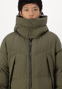 Olivově zelená péřová bunda s vysokým límcem, knoflíkovým zapínáním, zipem vpředu a prošívaným vzorem na hrudi a po stranách.