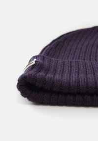 Bonnet en tricot violet, avec une texture côtelée et un revers replié. Petite étiquette en tissu avec logo attachée près du bord.