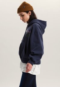 Marineblauw capuchontrui met voorkangoeroezak, met een meerkleurige tekstafbeelding. Gecombineerd met een wit gelaagd overhemd en donkere spijkerbroek.