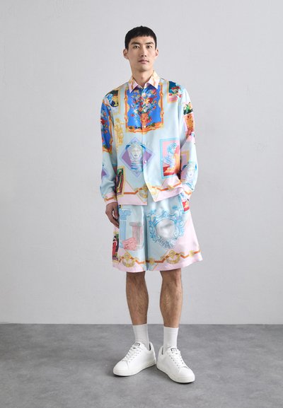 Camicetta a maniche corte, di colore azzurro chiaro, e pantaloni corti abbinati con motivi artistici colorati e dettagli dorati. Sneakers bianche completano il look.