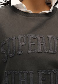 Ciemnoszara bluza z haftowanym napisem "SUPERDRY ATHLETE" w kontrastującym odcieniu. Miękka tkanina i widoczne ściągacze przy kołnierzu.