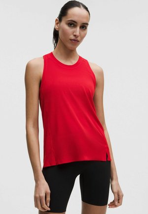 Femme aux longs cheveux bruns portant un débardeur de sport rouge et un cuissard noir, debout devant un fond clair uni.