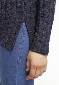 Marineblauwe geribbelde trui met zijsplitten, met een gestructureerd patroon. De mouwen lopen taps toe bij de pols en de trui wordt gedragen met blauwe jeans.