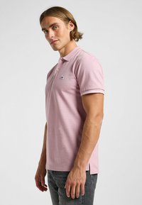 Polo shirt rose clair en coton avec col, deux boutons et bord blanc contrasté. Petit logo noir sur la poitrine.