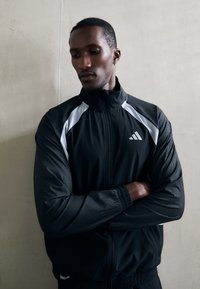 Black athletic jacket with white accents, featuring a high collar and textured sleeves. Adi logo on chest, elastic cuffs and hem.