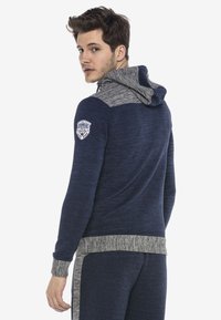 Marineblaues Kapuzensweatshirt mit grauer Kapuze, gerippten Bündchen und strukturiertem Stoff. Mit einem Logo-Patch auf der linken Schulter.