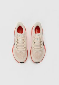 Bež mrežaste tenisice za trčanje s crvenim naglascima, okrugli vrh, crni swoosh logo i teksturirane vezice. Unutrašnjost ima kontrastnu crvenu podstavu.