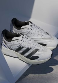 White athletic shoes with black stripes, textured surfaces, and a cushioned sole feature a streamlined design and contrasting accents.